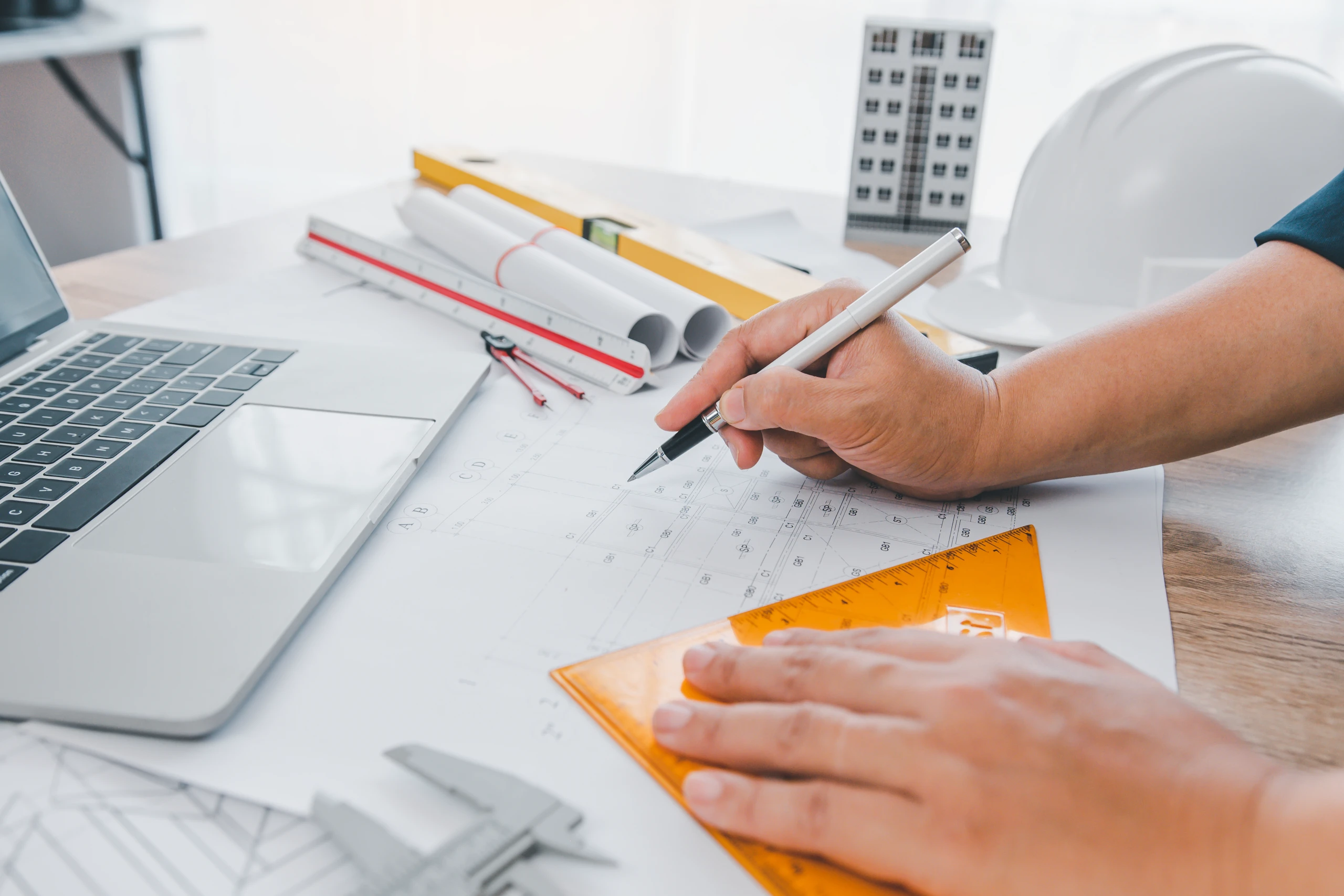 Architekt nanoszący pomiary ołówkiem na planie budowlanym przy laptopie i narzędziach kreślarskich
